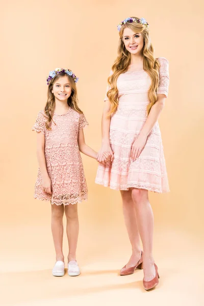 Beautiful woman with adorable daughter in elegant lacy dresses and floral wreaths holding hands on yellow background — Stock Photo