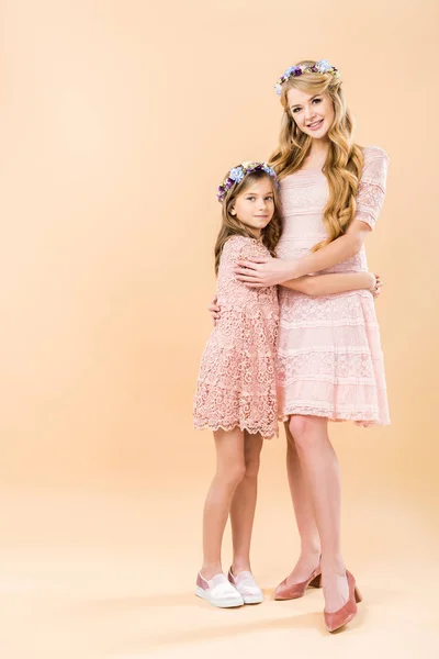Adorable hija y hermosa mamá en elegantes vestidos de encaje y coronas florales abrazándose y mirándose el uno al otro sobre fondo amarillo — Stock Photo
