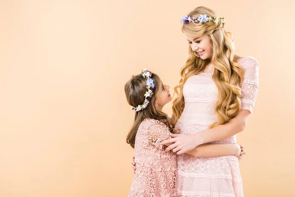 Mother and daughter in beautiful dresses and wreaths of flowers embracing and looking at each other on yellow background — Stock Photo