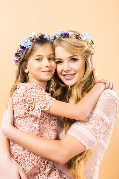 Mãe feliz e filha em coroas florais coloridas abraçando e olhando para a câmera no fundo amarelo — Fotografia de Stock