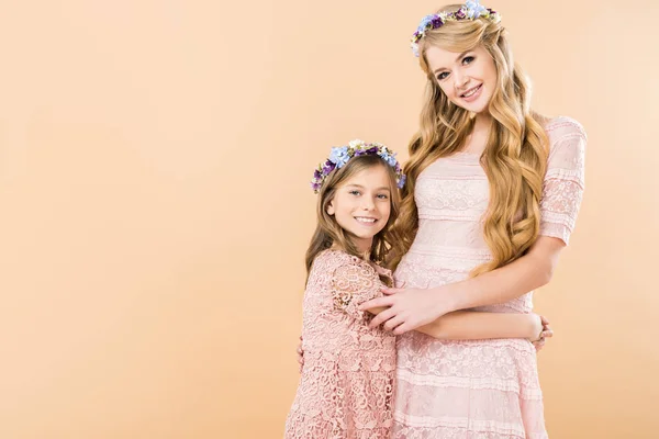Entzückende Tochter und schöne Mutter in eleganten Spitzenkleidern und bunten Blumenkränzen, die sich umarmen, während sie in die Kamera auf gelbem Hintergrund schauen — Stockfoto
