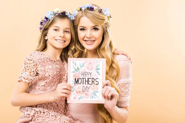 Sonrientes madre e hija en coronas florales sosteniendo feliz día de las madres tarjeta de felicitación sobre fondo amarillo - foto de stock
