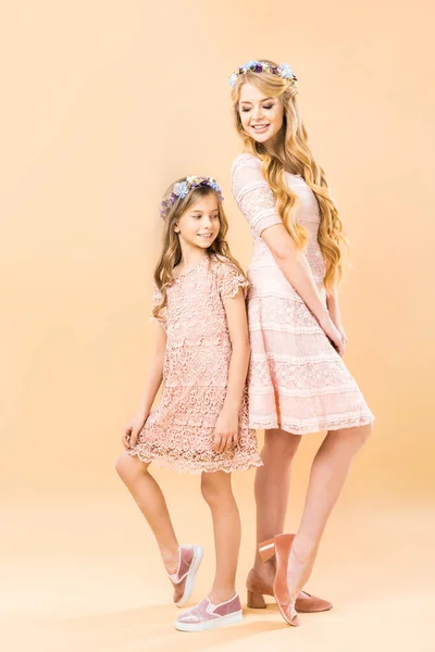 Мама и дочь в элегантных кружевных платьях и цветочных венках стоят спиной к спине и позируют перед камерой на желтом фоне — стоковое фото