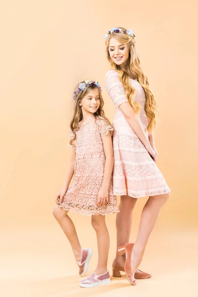 Mãe e filha em elegantes vestidos de renda e coroas florais de pé de volta para trás e posando para a câmera no fundo amarelo — Fotografia de Stock