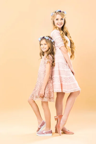 Hermosa mujer y linda hija en elegantes vestidos de encaje y coronas florales de pie espalda con espalda y posando en la cámara sobre fondo amarillo - foto de stock