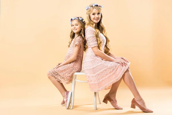 Happy mother and daughter in elegant lace dresses and floral wreaths sitting on same chair and looking at camera on yellow background — Stock Photo