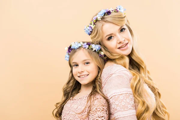 Mulher bonita e filha bonito em coroas florais coloridas de pé de volta para trás e olhando para a câmera no fundo amarelo — Fotografia de Stock