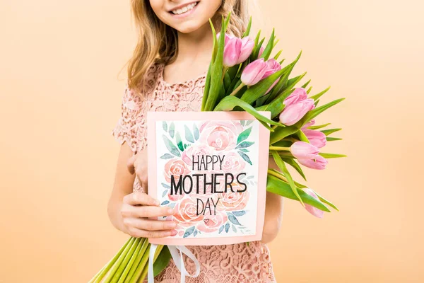 Vista recortada de niño sonriente sosteniendo feliz día de las madres tarjeta de felicitación y ramo de tulipanes rosados sobre fondo amarillo - foto de stock