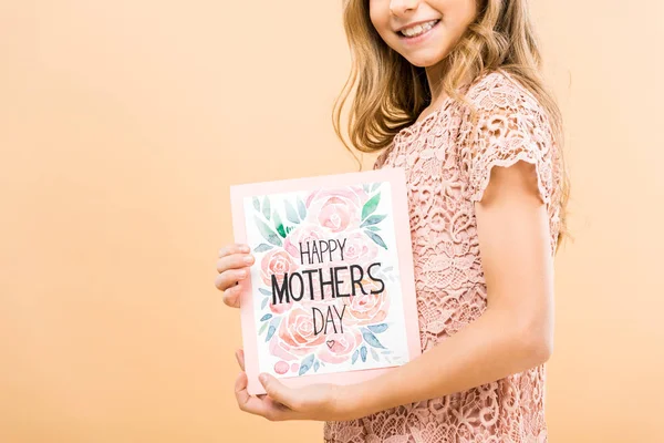 Vista parcial del niño sonriente en vestido de encaje rosa sosteniendo la tarjeta de felicitación feliz día de las madres sobre fondo amarillo - foto de stock