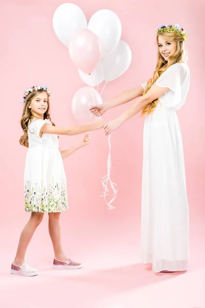 Beautiful mother and cute daughter in white elegant dresses and floral wreaths holding festive air balloons on pink background — Stock Photo