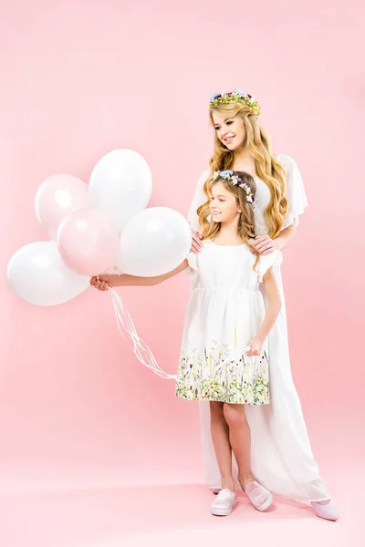 Niño lindo sosteniendo globos de aire festivos mientras está de pie cerca de la madre sonriente sobre fondo rosa - foto de stock