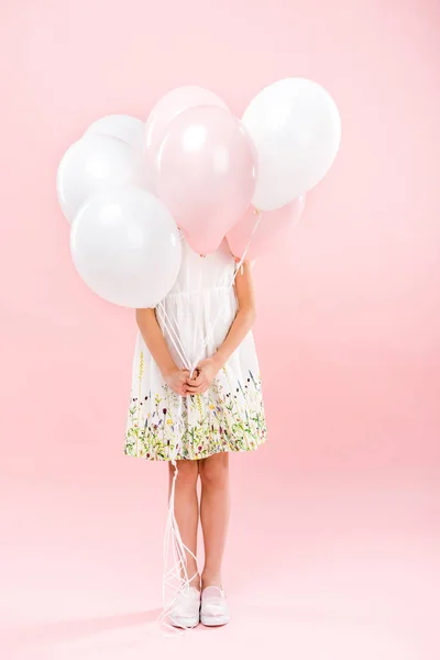 Child in delicate white dress hiding behind white and pink air balloons on pink background — Stock Photo