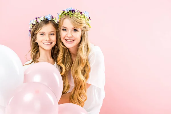 Mãe feliz e filha em coroas florais coloridas segurando balões de ar festivo no fundo rosa — Fotografia de Stock