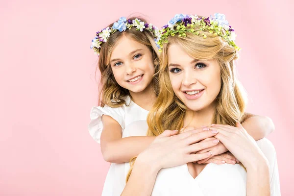 Pretty woman and cute child in colorful floral wreaths smiling and looking at camera on pink background — Stock Photo