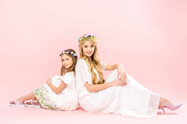 Atractiva mujer y linda hija en elegantes vestidos blancos y coronas florales sentados en el suelo espalda con espalda y mirando a la cámara en el fondo rosa - foto de stock