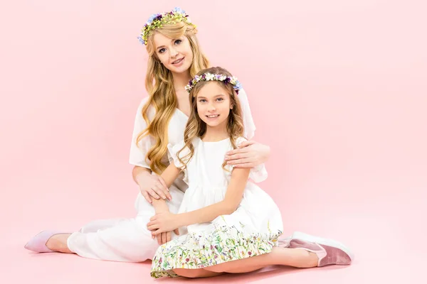 Hermosa mujer y adorable hija en elegantes vestidos blancos y coronas florales sentados en el suelo y mirando a la cámara en el fondo rosa - foto de stock