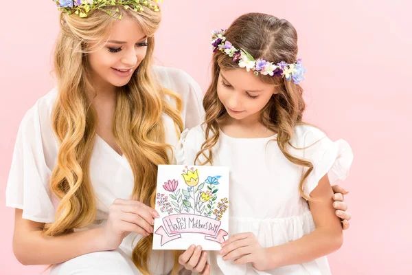 Hermosa madre e hija adorable en vestidos elegantes blancos y coronas florales con tarjeta de felicitación feliz día de las madres sobre fondo rosa - foto de stock
