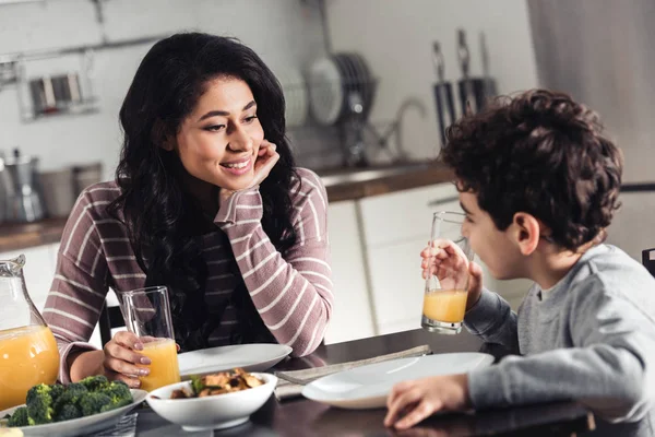 Glückliche lateinische Mutter schaut Sohn an, der ein Glas Orangensaft in der Küche hält — Stockfoto