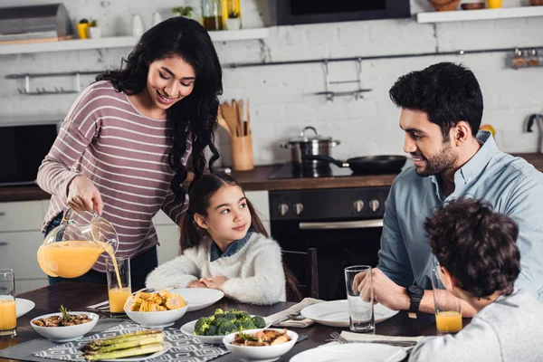 Fröhliche lateinamerikanische Mutter gießt während des Mittagessens Orangensaft in Glas von Tochter in der Nähe von Ehemann und Sohn — Stockfoto