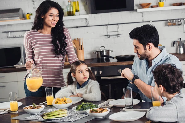 Cheerful latin mother holding jar with orange juice near daughter, husband and son during lunch — Stock Photo