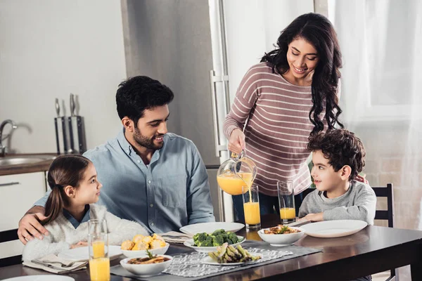 Joyeuse mère latine versant du jus d'orange dans un verre de mari heureux près de la fille et du fils pendant le déjeuner — Photo de stock