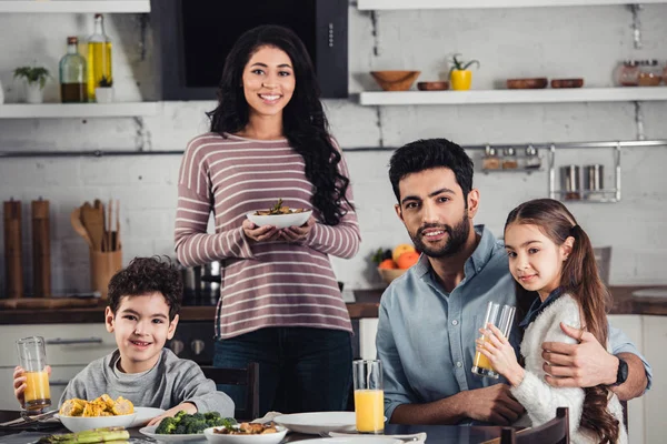 Mãe latina alegre segurando prato com comida perto de filho bonito, marido e filha durante o almoço — Fotografia de Stock