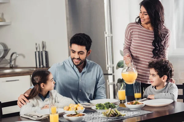 Fröhliche lateinische Mutter hält Glas mit Orangensaft in der Nähe ihres Sohnes, während sie den Ehemann beim Mittagessen dabei beobachtet, wie er Tochter umarmt — Stockfoto