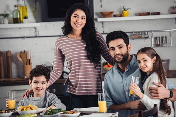 Cheerful latin mother hugging cute son and husband near daughter during lunch — Stock Photo