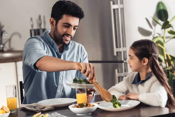 Cheerful latin father putting broccoli in plate of cute daughter at home — Stock Photo