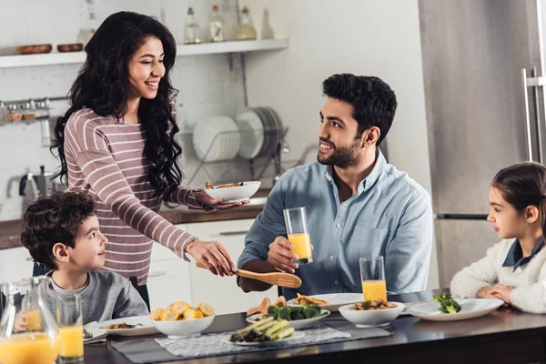 Fröhliche lateinische Frau schaut Ehemann an, während sie beim Mittagessen Teller in der Nähe von Tochter und Sohn hält — Stockfoto