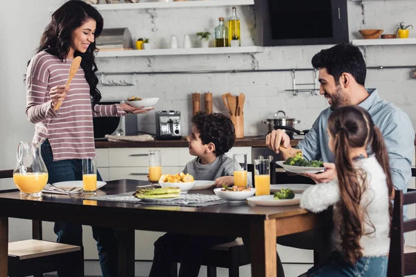 Attractive latin woman looking at son while holding plate near daughter and husband during lunch — Stock Photo