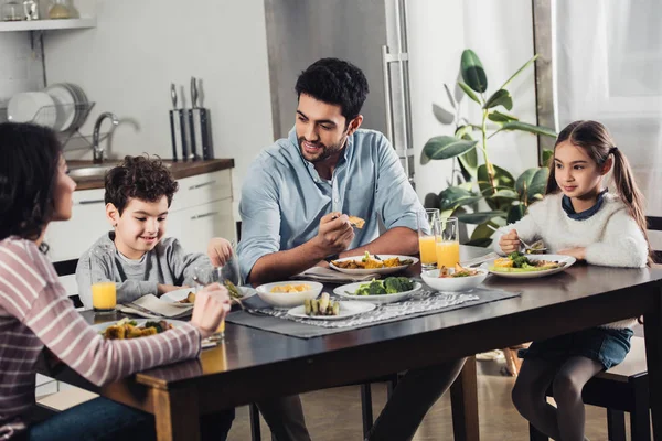 Guapo padre latino mirando hijo mientras almorzando cerca de hija y esposa - foto de stock