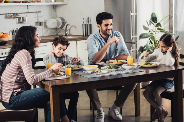 Feliz madre latina mirando linda hija almorzando cerca de padre e hijo en casa - foto de stock