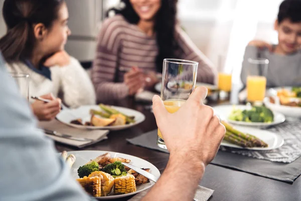 Foco seletivo de vidro com suco de laranja na mão do homem almoçando com a família latina em casa — Fotografia de Stock