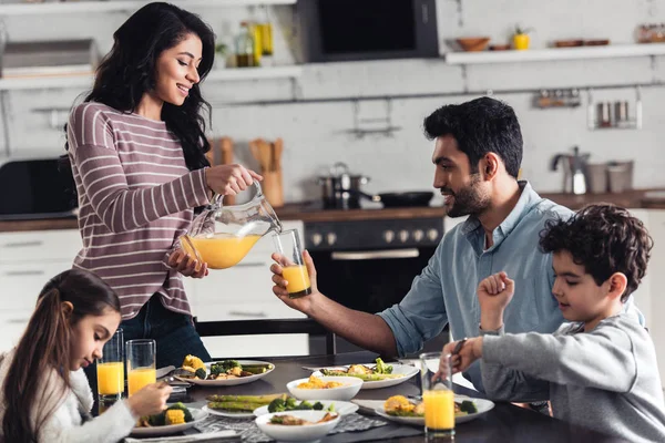 Happy latin mother smiling while pouring orange juice during lunch near hispanic family — Stock Photo