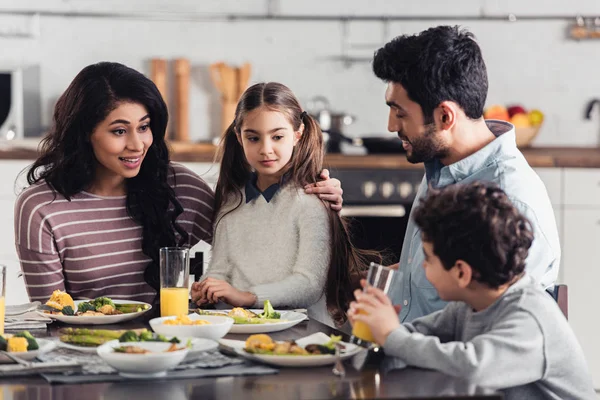 Attraktive lateinische Mutter umarmt Tochter, während sie ihren Sohn in der Nähe des Ehemannes zu Hause ansieht — Stockfoto