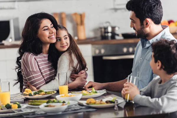 Cheerful latin woman hugging cute daughter while looking at husband near son at home — Stock Photo