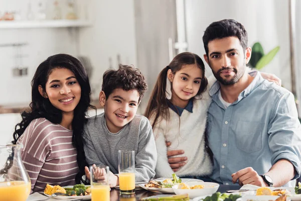 Cheerful latin family hugging and looking at camera during lunch at home — Stock Photo