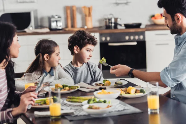 Cute latin kids looking at broccoli on fork in hand of father near mother at home — Stock Photo