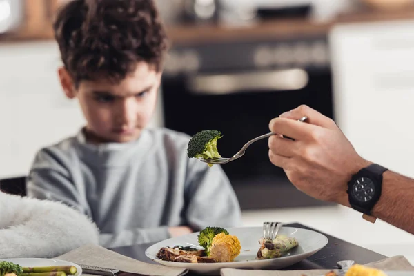 Selective focus of fork with green broccoli in hand of father with upset latin kid on background — Stock Photo