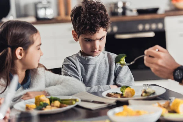 Selective focus of fork with green broccoli in hand of father with upset latin boy near sister on background — Stock Photo