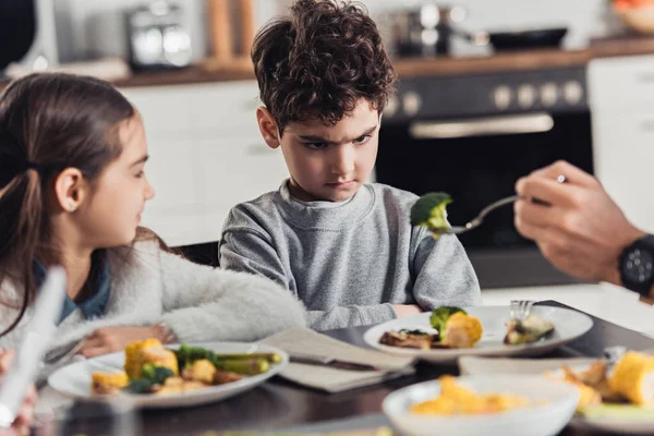 Foyer sélectif de fourchette avec brocoli vert dans la main du père avec garçon latino bouleversé près de soeur sur fond — Photo de stock