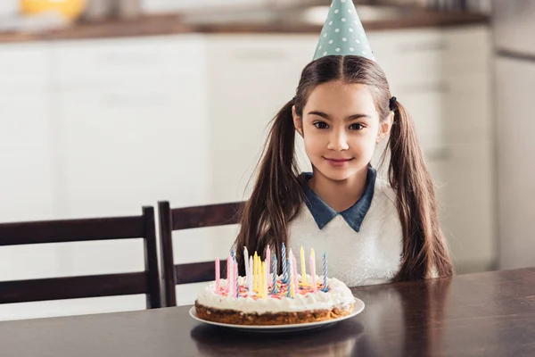 Niño latino alegre en gorra de fiesta sentado cerca de pastel de cumpleaños con velas en casa - foto de stock