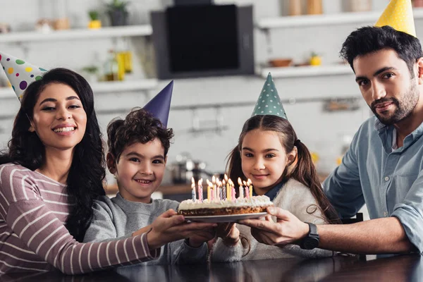 Alegre familia hispana sosteniendo pastel de cumpleaños y sonriendo en casa - foto de stock