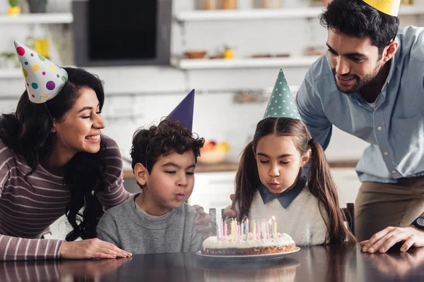 Crianças hispânicas bonitos soprando em velas no bolo de aniversário perto de pai feliz e mãe em casa — Fotografia de Stock