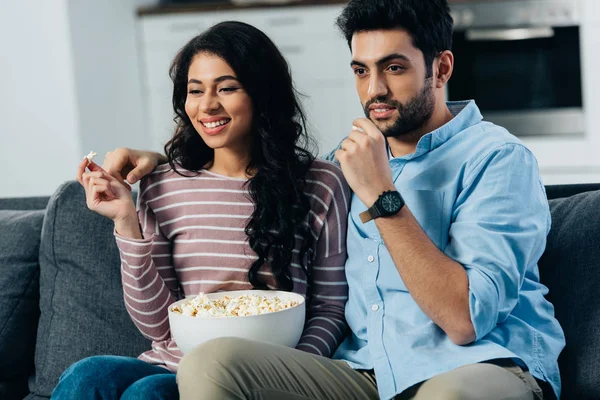 Feliz mulher latina segurando pipocas perto bonito marido enquanto assiste tv em casa — Fotografia de Stock