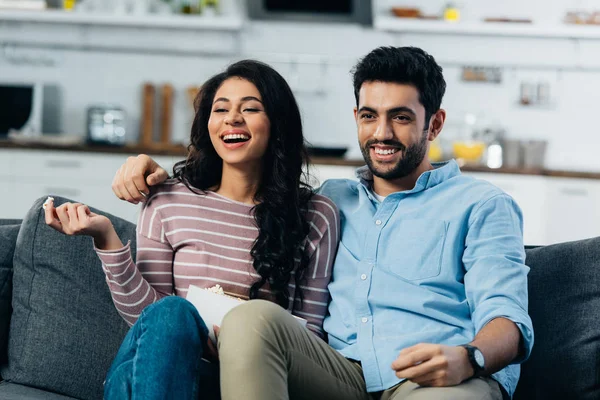 Feliz pareja latina viendo la televisión en casa con un tazón de palomitas de maíz - foto de stock