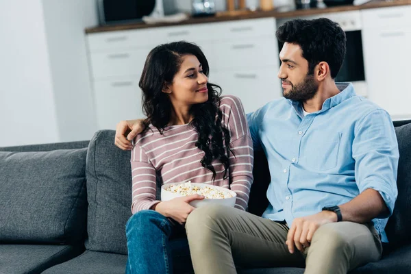 Atractiva mujer latina sosteniendo tazón con palomitas de maíz y mirando al marido en casa - foto de stock