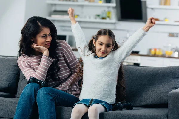 Mignon latin enfant mettre les mains au-dessus de la tête après avoir joué jeu vidéo près bouleversé mère à la maison — Photo de stock