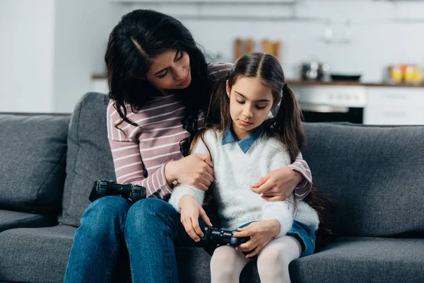 Mère latine embrassant fille bouleversée après avoir joué à un jeu vidéo à la maison — Photo de stock